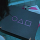 The Squid Game logo printed on a box, very similar to the distinctive PlayStation shapes.